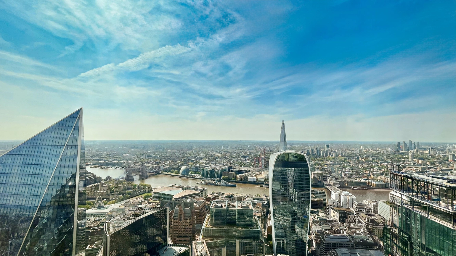 A high-angle view of London under clear blue skies. There is a mix of modern skyscrapers with reflective glass facades, like the Fenchurch Building (also known as The Walkie-Talkie). The River Thames winds through the photograph. A high-angle view of London under clear blue skies. There is a mix of modern skyscrapers with reflective glass facades, like the Fenchurch Building (also known as The Walkie-Talkie). The River Thames winds through the photograph.
