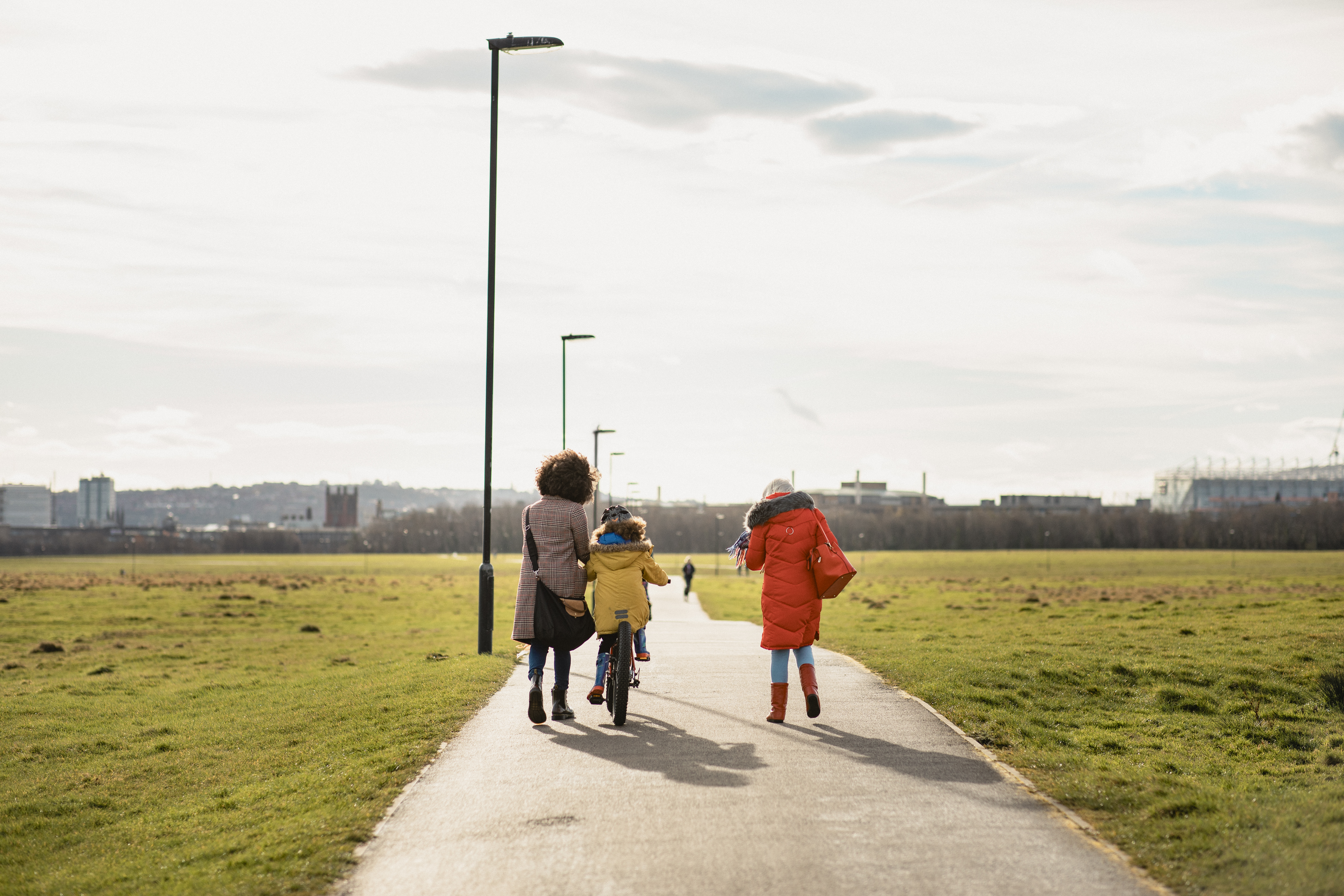 A wide paved path runs through a large green park under a bright, partly cloudy sky. Three people are walking away from the camera along the path: one person is walking with a child riding a bicycle, and another person is walking beside them. They are all wearing colourful winter coats. Tall streetlights line the path, and in the distance, there is a city skyline with low-rise buildings and a large stadium-like structure on the right.