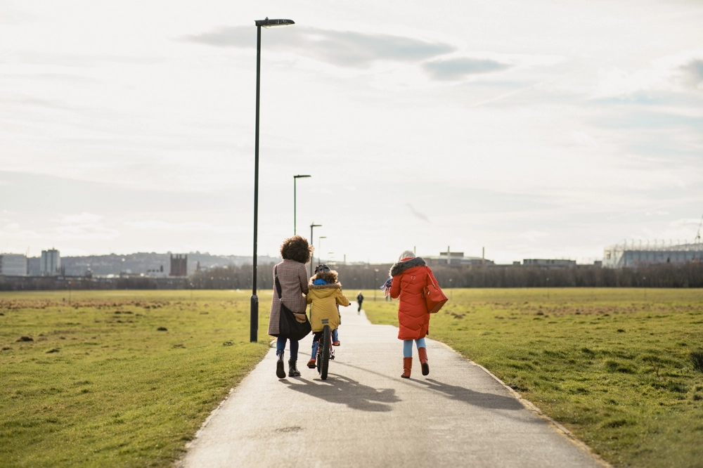 A wide paved path runs through a large green park under a bright, partly cloudy sky. Three people are walking away from the camera along the path: one person is walking with a child riding a bicycle, and another person is walking beside them. They are all wearing colourful winter coats. Tall streetlights line the path, and in the distance, there is a city skyline with low-rise buildings and a large stadium-like structure on the right. A wide paved path runs through a large green park under a bright, partly cloudy sky. Three people are walking away from the camera along the path: one person is walking with a child riding a bicycle, and another person is walking beside them. They are all wearing colourful winter coats. Tall streetlights line the path, and in the distance, there is a city skyline with low-rise buildings and a large stadium-like structure on the right.