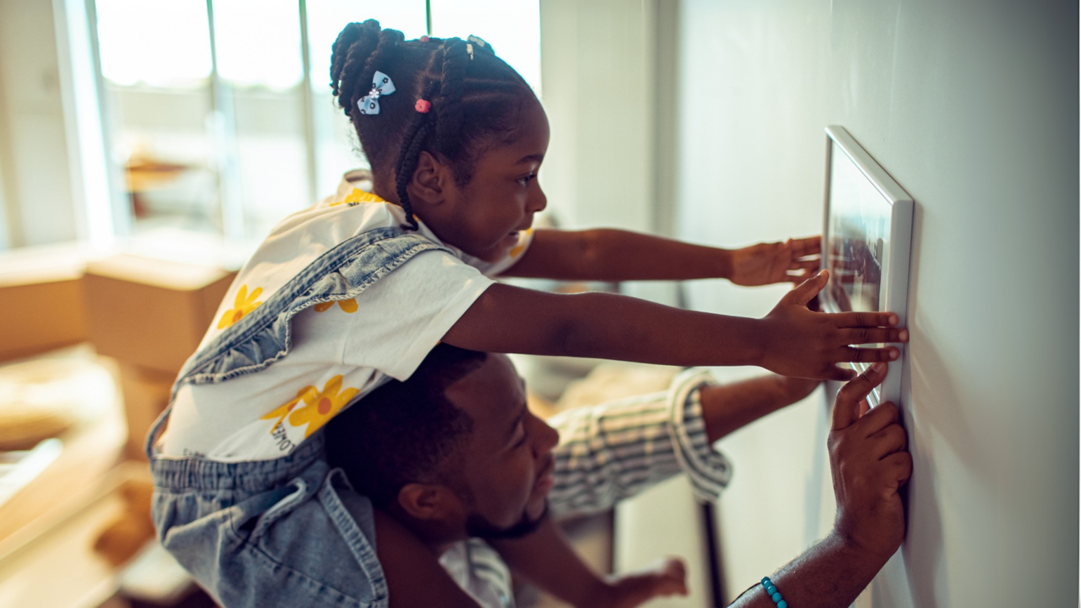 A child sits on an adult’s shoulders as they hang a picture frame on the wall of their new home. A child sits on an adult’s shoulders as they hang a picture frame on the wall of their new home.