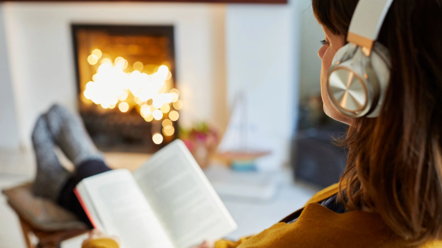 Female reading her book and listening to the music through headset in front of fire place in their living room Female reading her book and listening to the music through headset in front of fire place in their living room