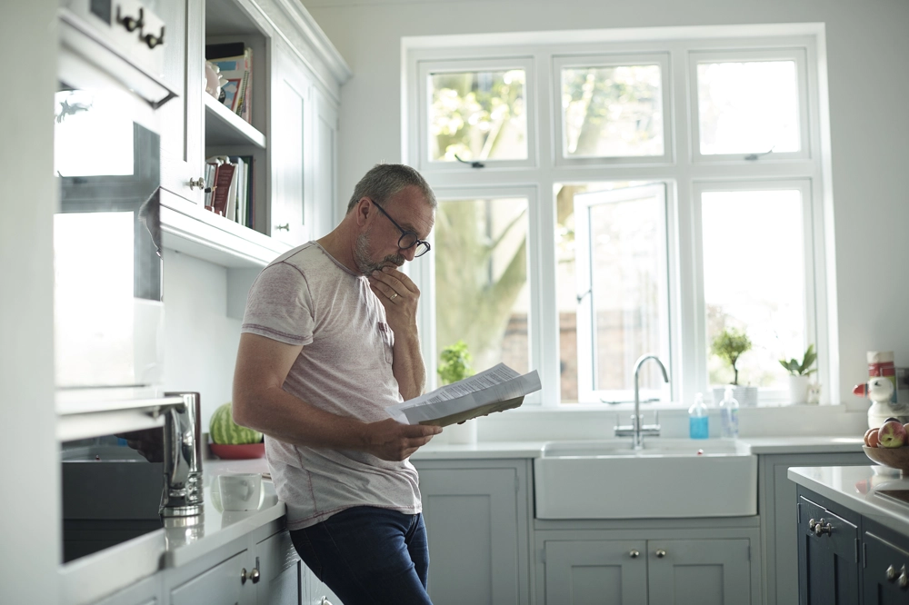 A person standing in a bright, modern kitchen, leaning against the counter while reading a document. Sunlight comes through large windows above the farmhouse-style sink, illuminating the light grey cabinets and countertops. A person standing in a bright, modern kitchen, leaning against the counter while reading a document. Sunlight comes through large windows above the farmhouse-style sink, illuminating the light grey cabinets and countertops.