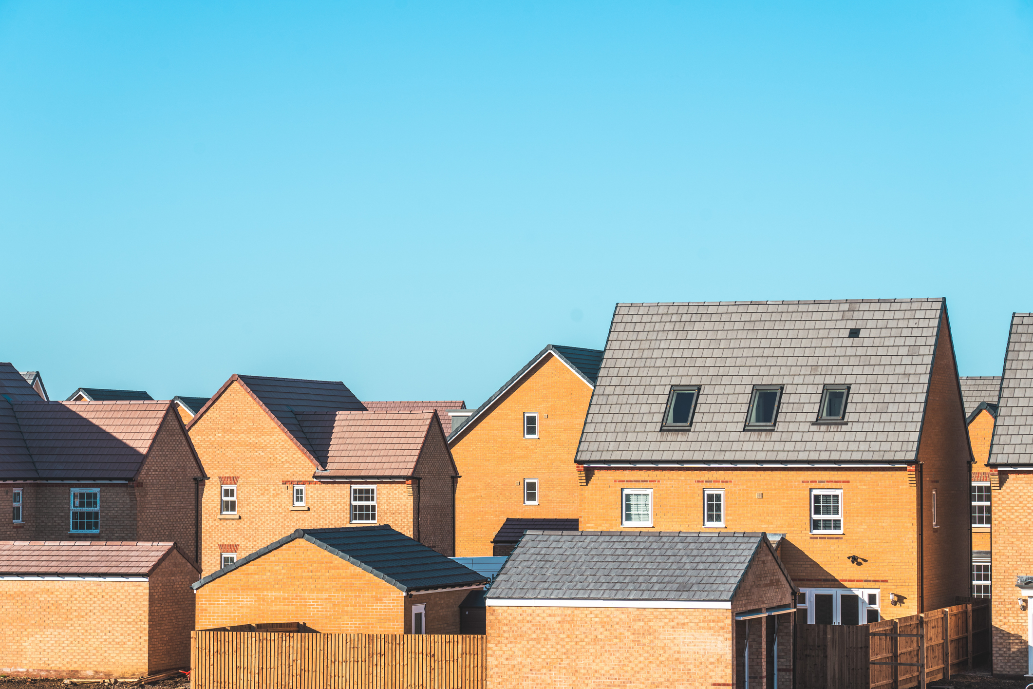Modern new‑build family houses built in yellow brick with dark pitched tiled roofs under a clear blue daytime sky.