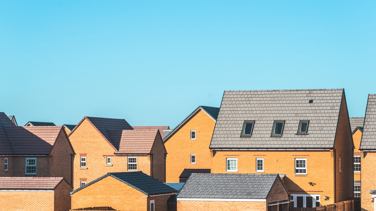 Modern new‑build family houses built in yellow brick with dark pitched tiled roofs under a clear blue daytime sky.