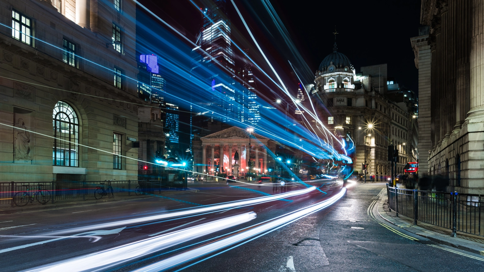 Night view of the Bank Junction in London, UK, with illuminated buildings and skyscrapers in the background. Bright streetlights create starburst effects, and light trails from passing vehicles flow across the foreground. Night view of the Bank Junction in London, UK, with illuminated buildings and skyscrapers in the background. Bright streetlights create starburst effects, and light trails from passing vehicles flow across the foreground.
