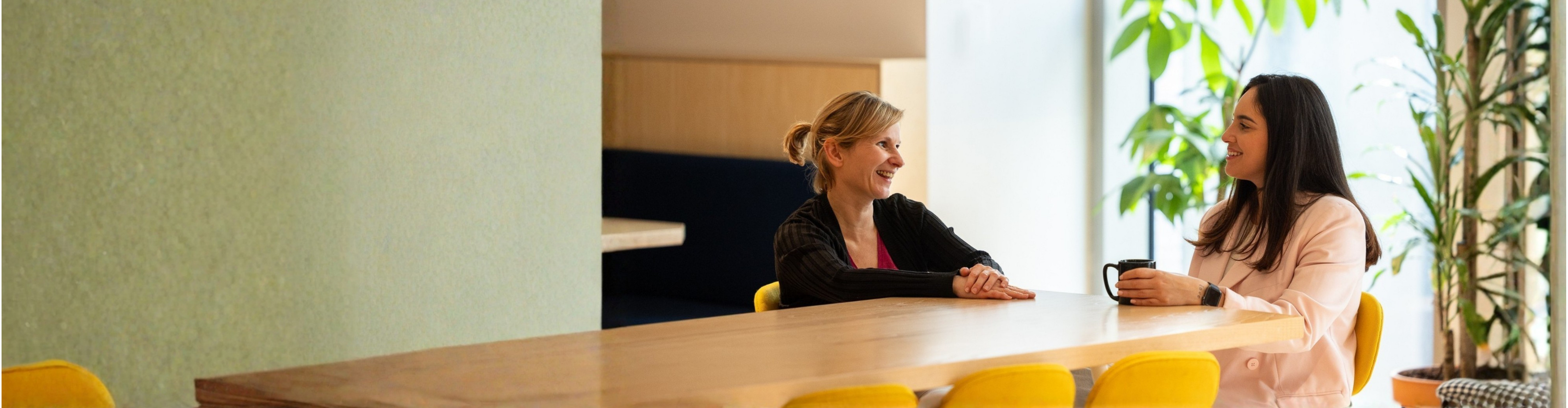 Two colleagues seated at a wooden table with yellow chairs in a bright, plant-filled room with large windows. The person on the left wears a dark jacket, while the one on the right wears a light blazer and holds a black mug. They appear to be engaged in a conversation.