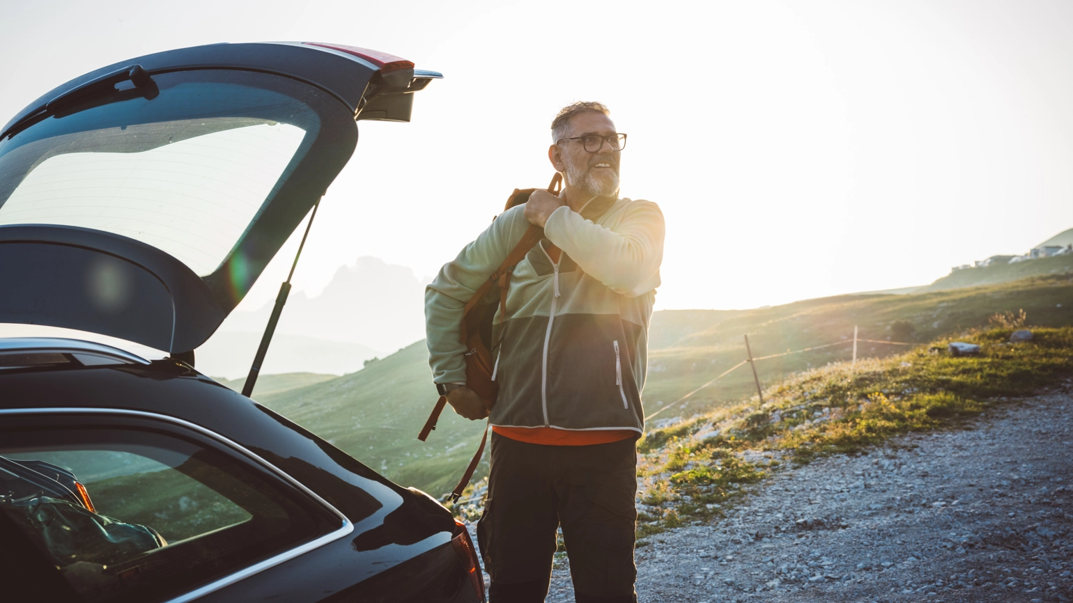 Person standing beside an open car trunk on a gravel path, adjusting a backpack strap. The scene features grassy slopes and distant mountain peaks, illuminated by a warm setting sunlight. Person standing beside an open car trunk on a gravel path, adjusting a backpack strap. The scene features grassy slopes and distant mountain peaks, illuminated by a warm setting sunlight.