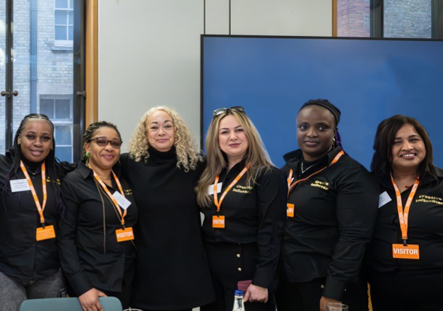 Six people stand together indoors, all wearing black clothing and orange visitor badges.