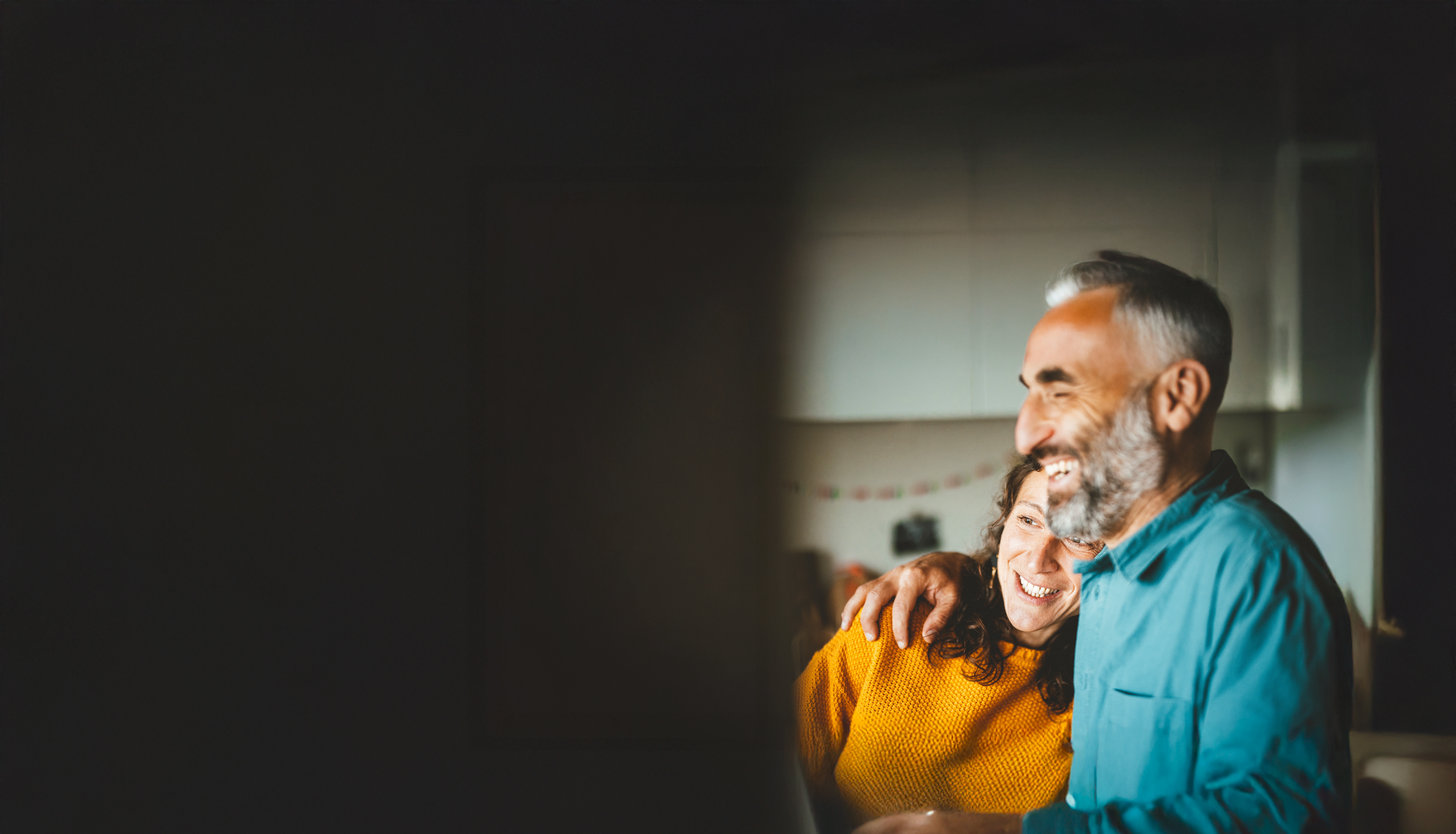 A person in a teal shirt with an arm around another person wearing a yellow sweater inside a softly lit kitchen.