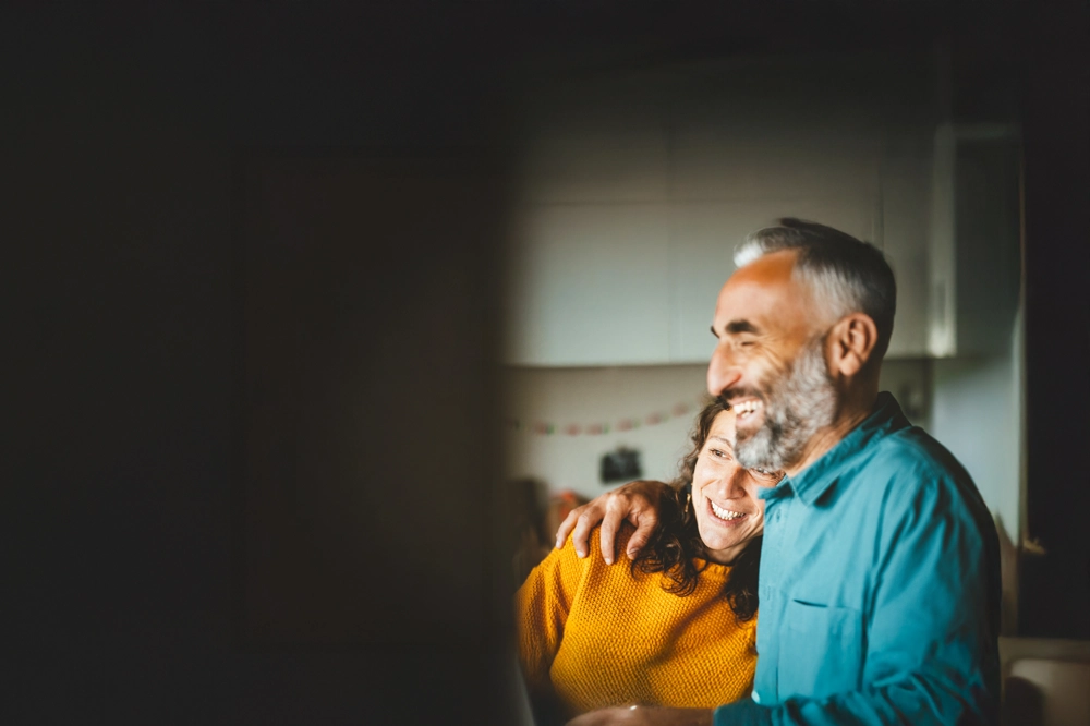 A person in a teal shirt with an arm around another person wearing a yellow sweater inside a softly lit kitchen. A person in a teal shirt with an arm around another person wearing a yellow sweater inside a softly lit kitchen.