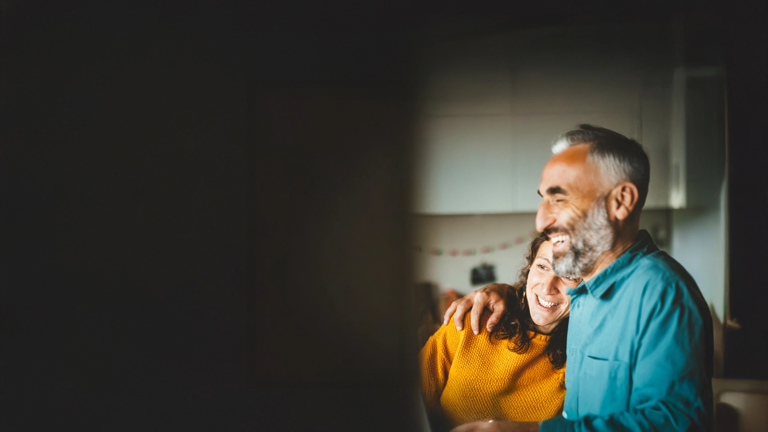 A person in a teal shirt with an arm around another person wearing a yellow sweater inside a softly lit kitchen. A person in a teal shirt with an arm around another person wearing a yellow sweater inside a softly lit kitchen.