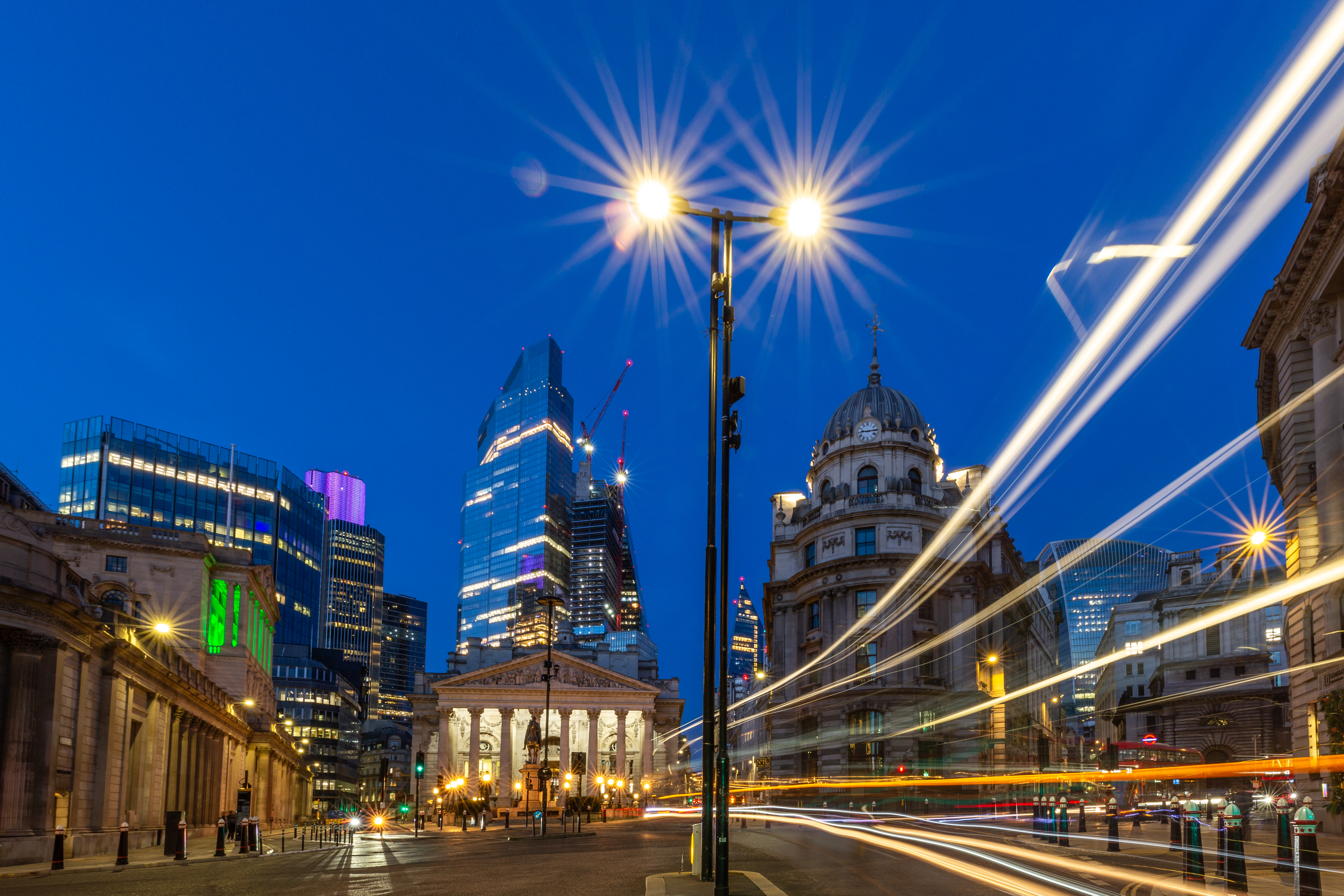 Night view of the Bank Junction in London, UK, with illuminated buildings and skyscrapers in the background. Bright streetlights create starburst effects, and light trails from passing vehicles flow across the foreground.