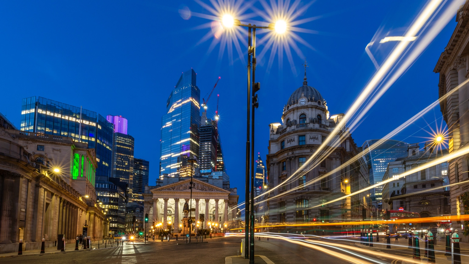 Night view of the Bank Junction in London, UK, with illuminated buildings and skyscrapers in the background. Bright streetlights create starburst effects, and light trails from passing vehicles flow across the foreground. Night view of the Bank Junction in London, UK, with illuminated buildings and skyscrapers in the background. Bright streetlights create starburst effects, and light trails from passing vehicles flow across the foreground.