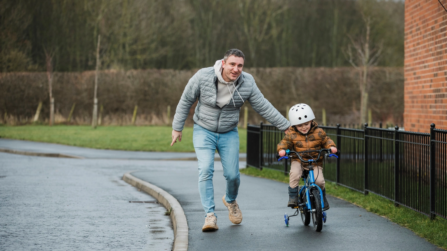 A father rests a reassuring hand on his young son's back as he learns to ride his bike on the footpath outside of his home in the North East of England. The young boy is smiling as he controls the bike on the footpath. A father rests a reassuring hand on his young son's back as he learns to ride his bike on the footpath outside of his home in the North East of England. The young boy is smiling as he controls the bike on the footpath.