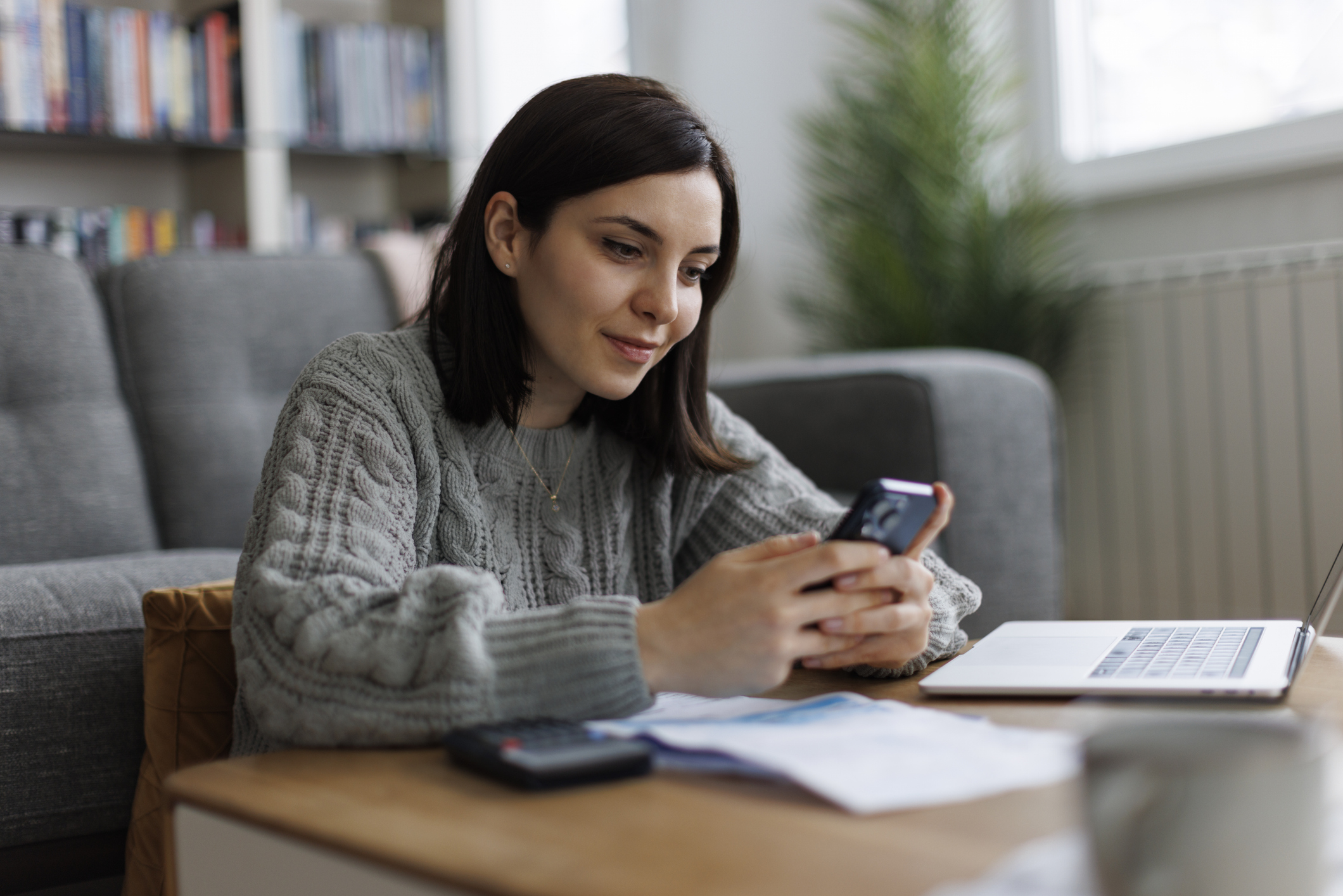 A person leaning on a wooden coffee table in a living room, wearing a grey knitted jumper and holding a smartphone. On the coffee table are scattered papers, a calculator, and an open laptop. In the background, there is a grey sofa, a tall green plant, and shelves filled with books. Daylight comes through a window on the right side.