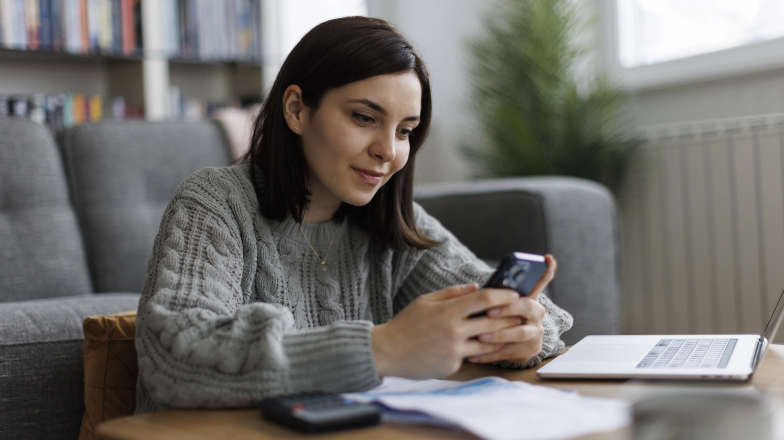 A person leaning on a wooden coffee table in a living room, wearing a grey knitted jumper and holding a smartphone. On the coffee table are scattered papers, a calculator, and an open laptop. In the background, there is a grey sofa, a tall green plant, and shelves filled with books. Daylight comes through a window on the right side. A person leaning on a wooden coffee table in a living room, wearing a grey knitted jumper and holding a smartphone. On the coffee table are scattered papers, a calculator, and an open laptop. In the background, there is a grey sofa, a tall green plant, and shelves filled with books. Daylight comes through a window on the right side.