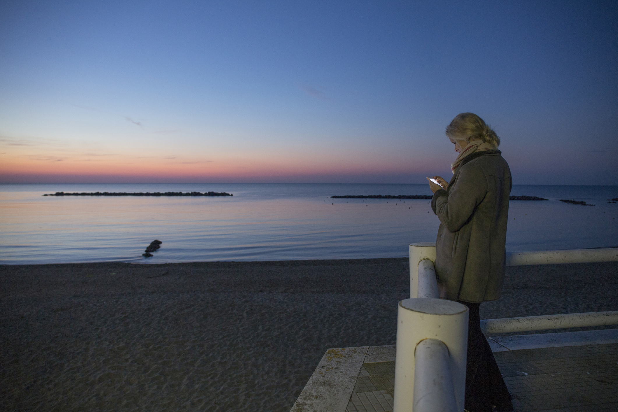 A person wearing a winter coat standing on a seaside promenade at dusk, illuminated by the light of their phone while facing a calm ocean and a sky that transitions from deep blue to soft pink near the horizon.