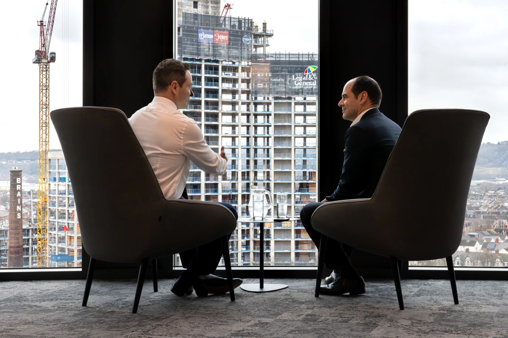 António Simões, CEO of Legal & General, seated by a window with a colleague, with construction workers visible outside. António Simões, CEO of Legal & General, seated by a window with a colleague, with construction workers visible outside.