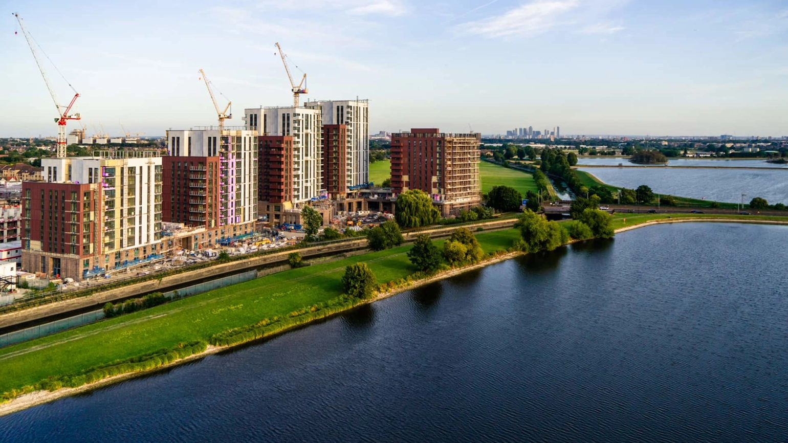 Aerial view of the New Acres scheme in Wandsworth, London. The image captures a vibrant new community taking shape, with contemporary mid-rise residential buildings under construction and cranes positioned across the site. The scheme is set alongside expansive water and green spaces, creating a balance of urban living and natural surroundings. In the distance, the London skyline is visible under a clear blue sky. Aerial view of the New Acres scheme in Wandsworth, London. The image captures a vibrant new community taking shape, with contemporary mid-rise residential buildings under construction and cranes positioned across the site. The scheme is set alongside expansive water and green spaces, creating a balance of urban living and natural surroundings. In the distance, the London skyline is visible under a clear blue sky.