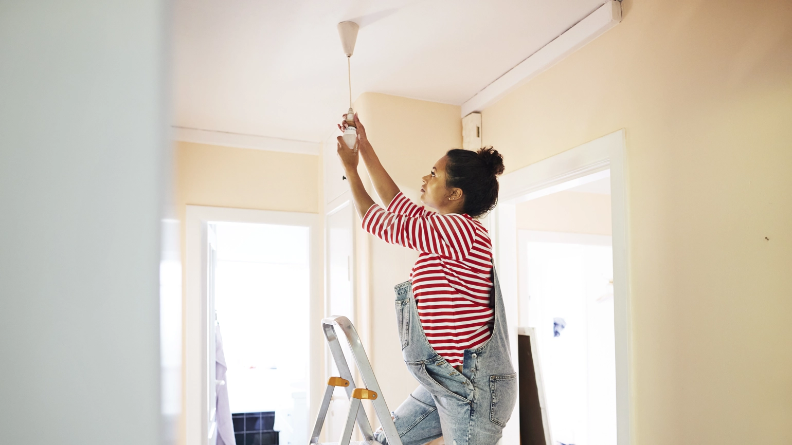 A pregnant woman changing a light bulb on a corridor ceiling while standing on ladder. A pregnant woman changing a light bulb on a corridor ceiling while standing on ladder.