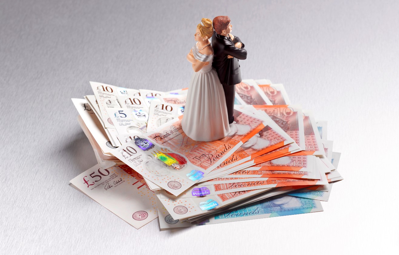 Wedding cake topper of a bride and groom standing back-to-back with arms crossed, placed on a stack of banknotes.