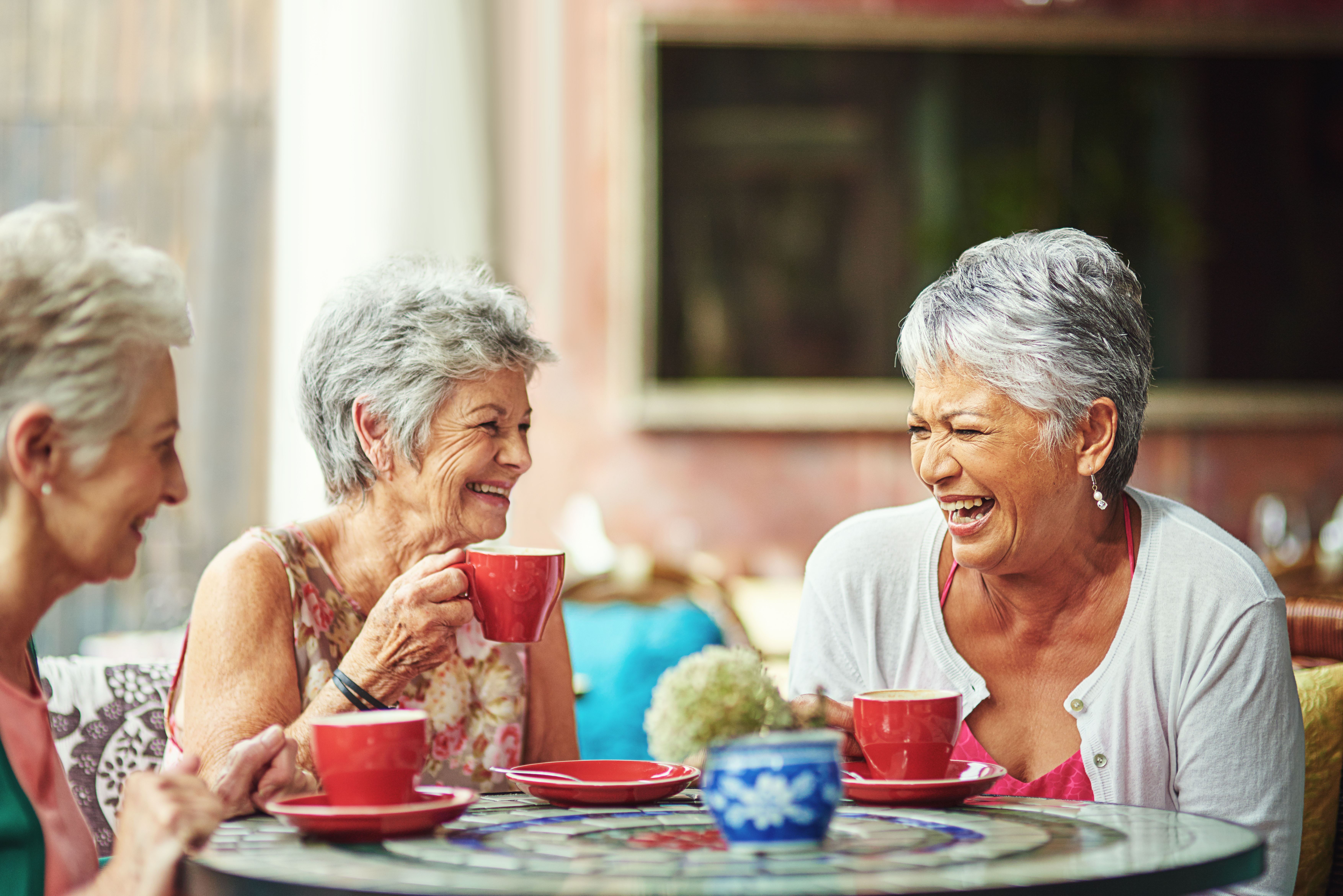 Three adults sitting at a café table, drinking from red cups and talking around a colourful mosaic table.