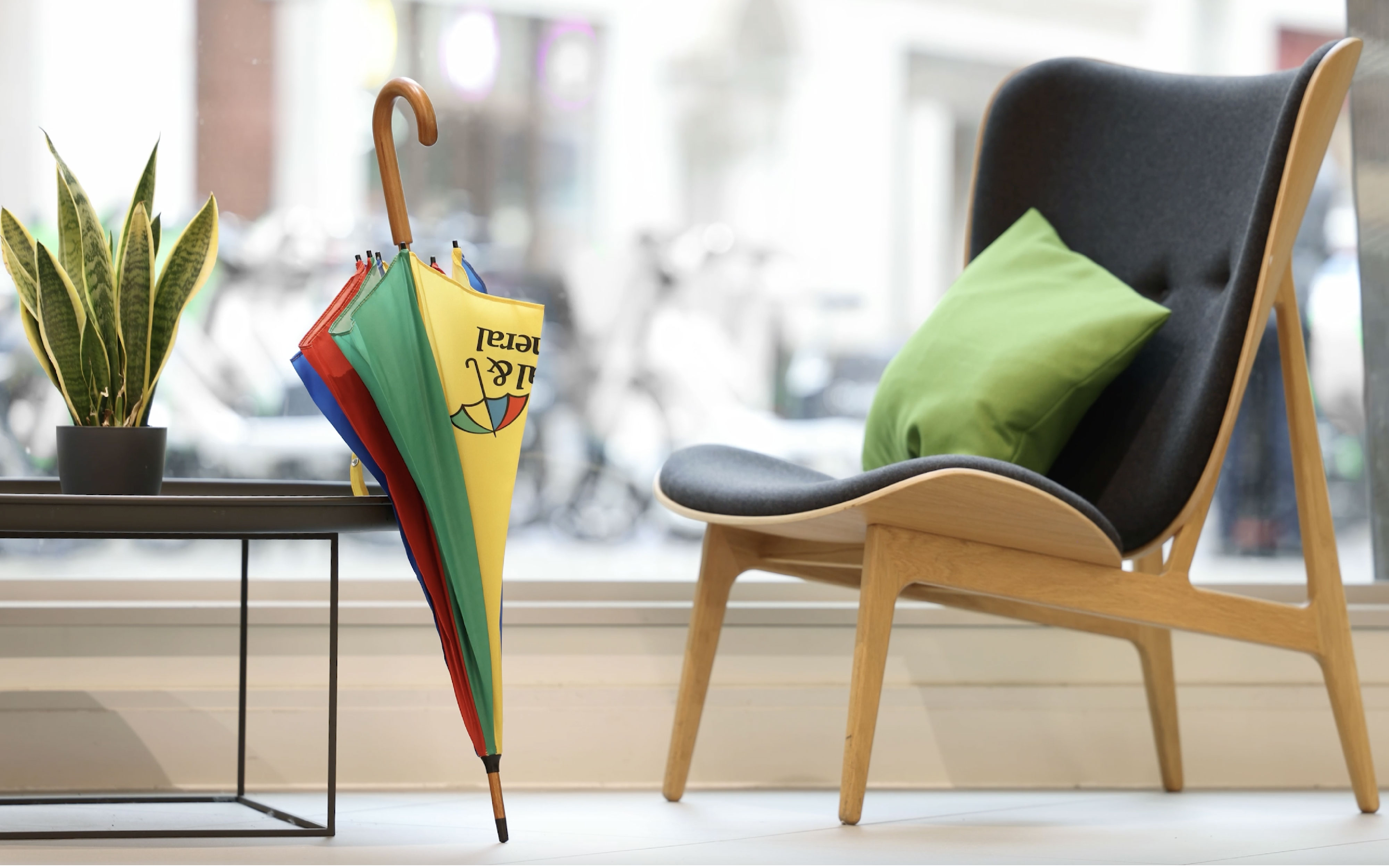 A wooden chair with dark upholstery and a green cushion in front of a large window. Next to the chair is a small black table with a potted plant. On the floor, leaning against the table, is a colourful umbrella with yellow, green, red, and blue panels. The Legal & General logo is visible on the yellow panel.