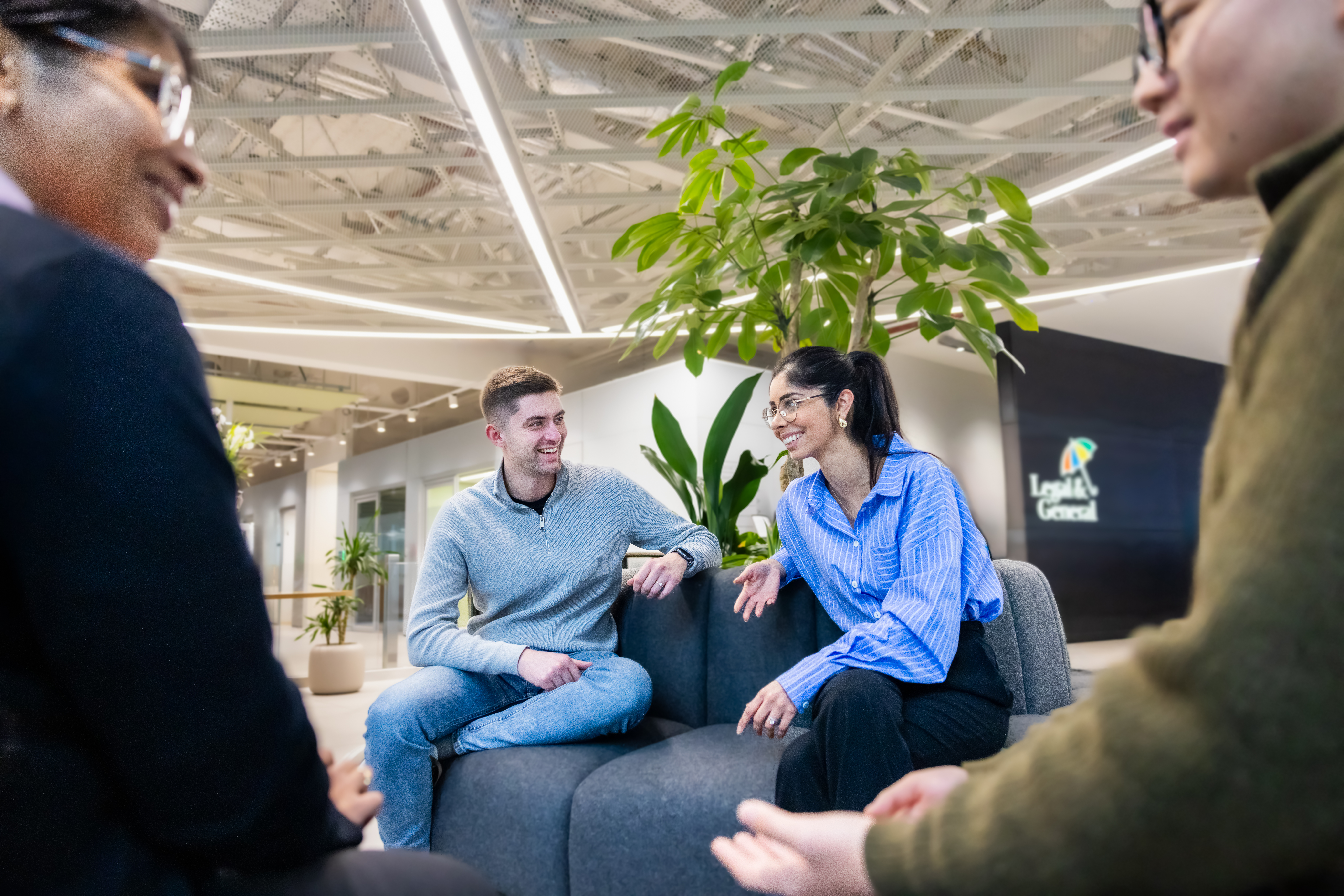 Four people seated on a dark grey circular sofa in Legal & General’s London office. Plants are arranged in the centre of the seating area, behind the people.